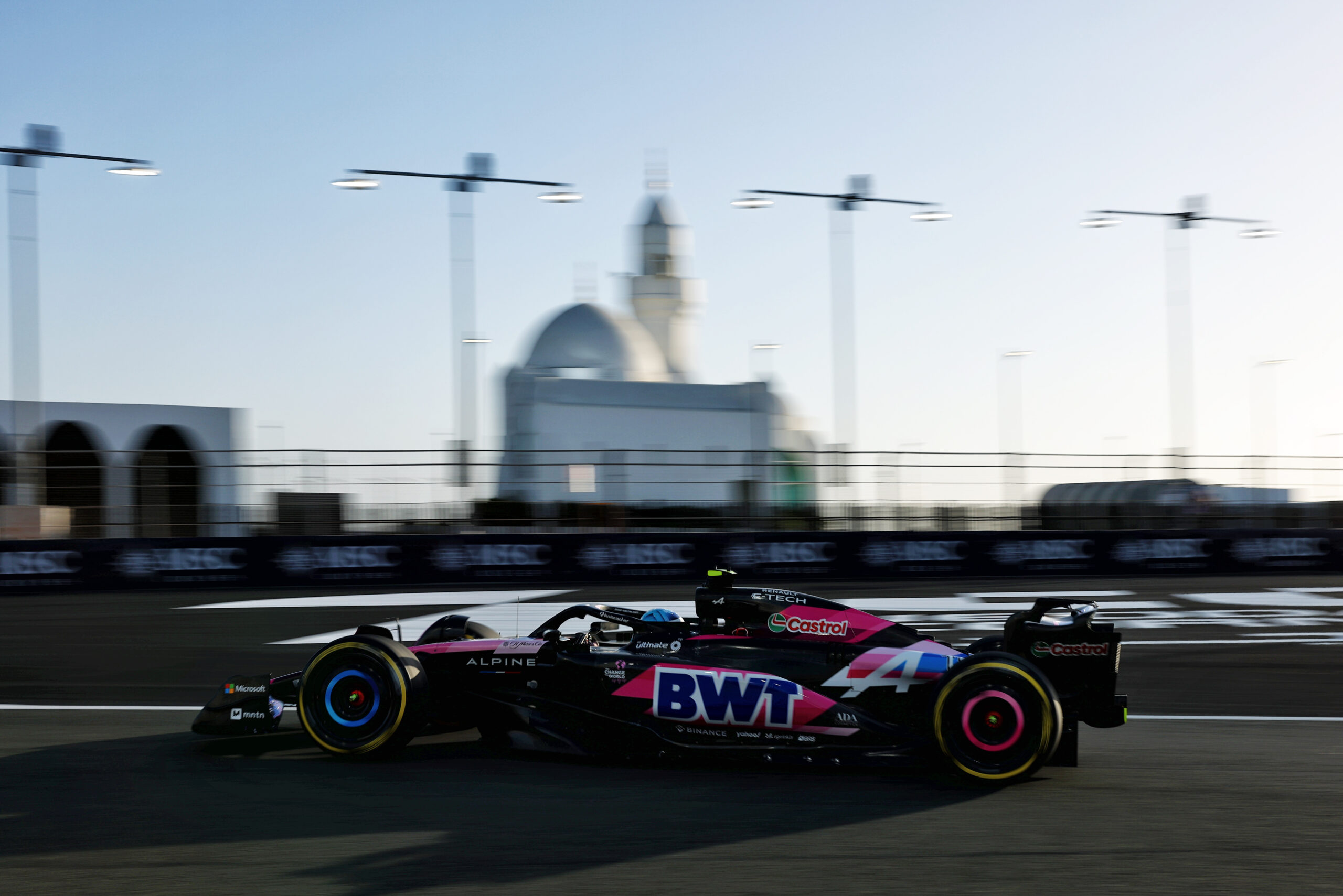 A Formula 1 car with BWT branding races on a track with a mosque and streetlights visible in the background. The scene is captured during daylight.