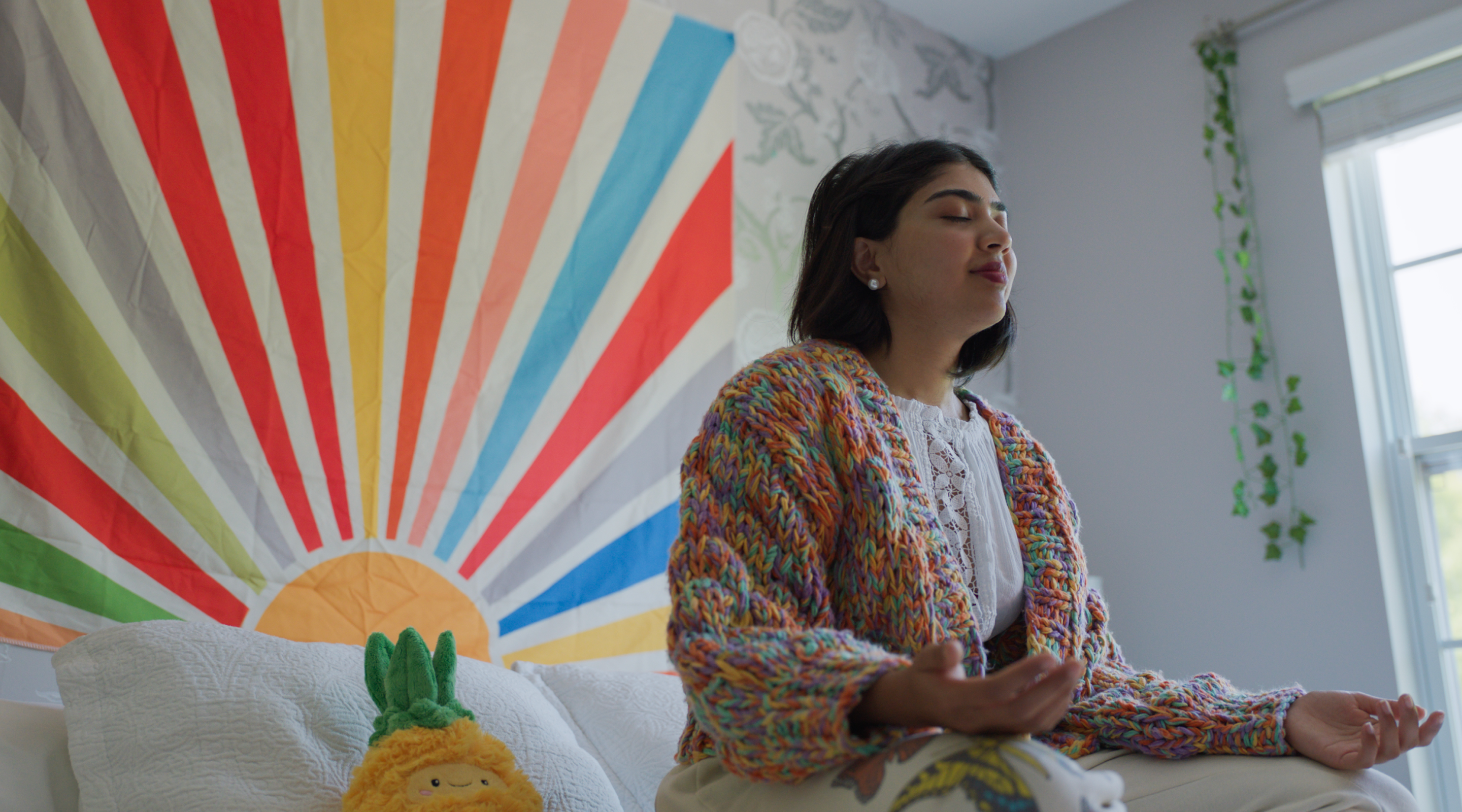Photo of girl meditating in her room with colorful rainbow tapestry behind her