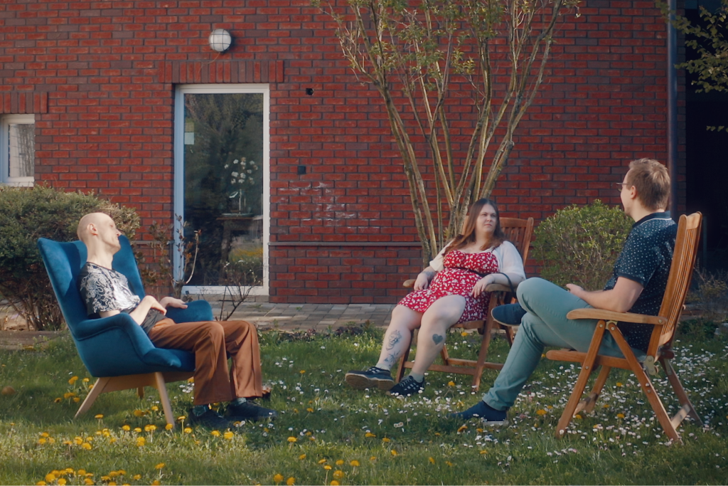 Two men and a woman sit in chairs in the grass outside of a building.