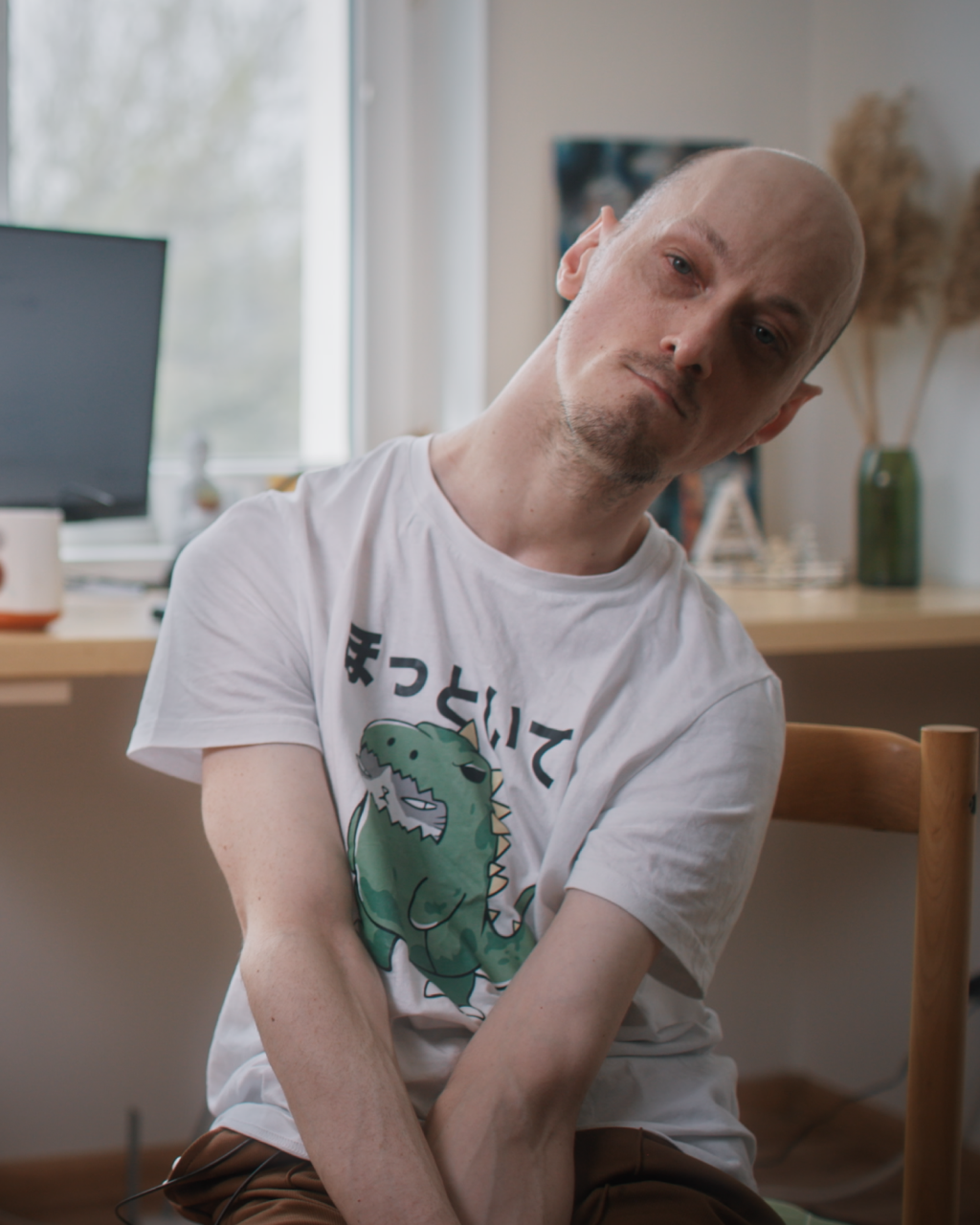 Portrait of a man in a chair, looking towards the camera. He wears a t-shirt with a cat dressed up as a dinosaur.