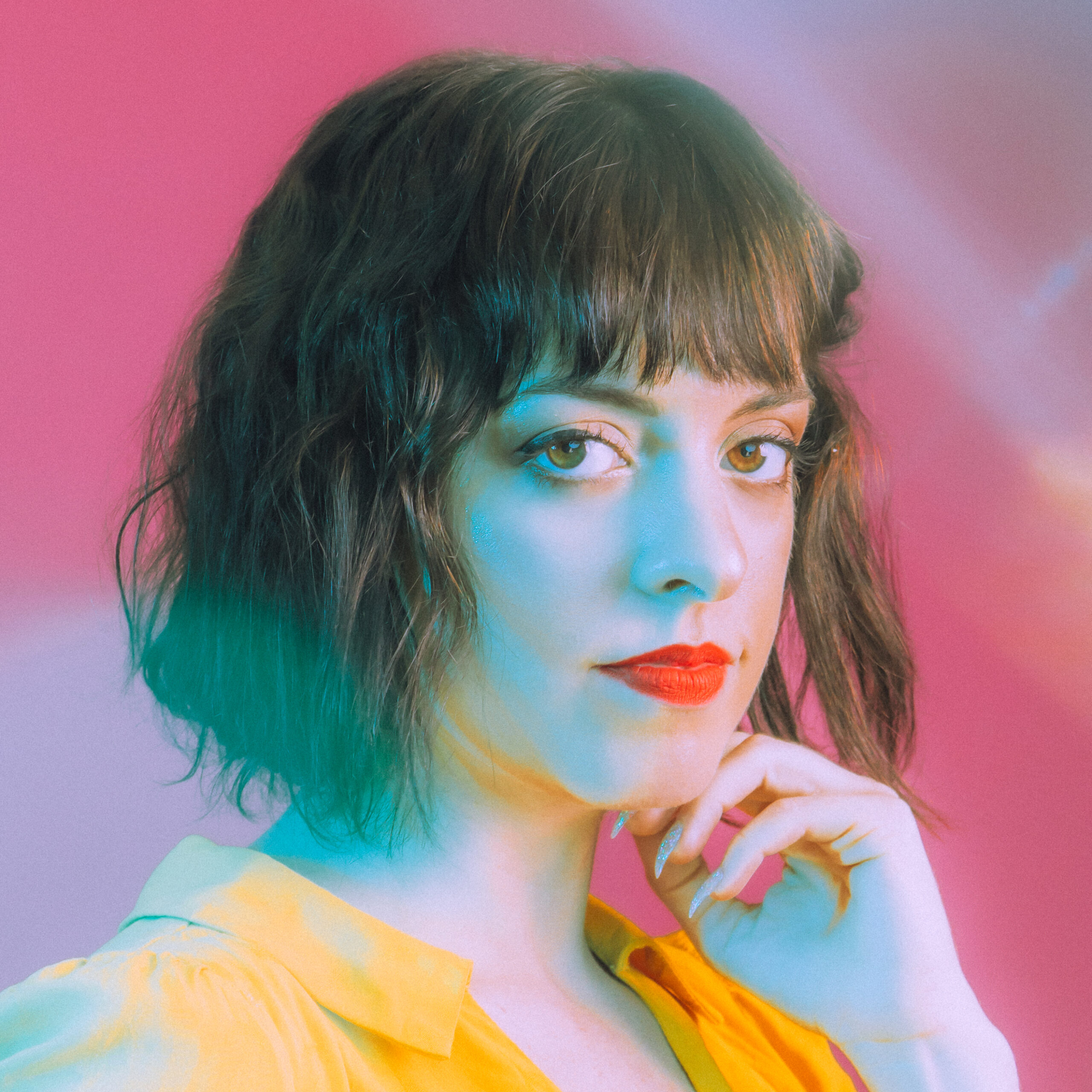 A vibrant portrait of an individual featuring dark short hair and bangs. The person is dressed in a yellow top and has her hand to her chin. The backdrop is a gradient pink.