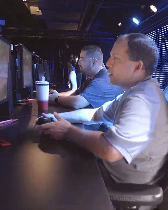 Two men seated with Xbox controllers in hand looking toward screen.