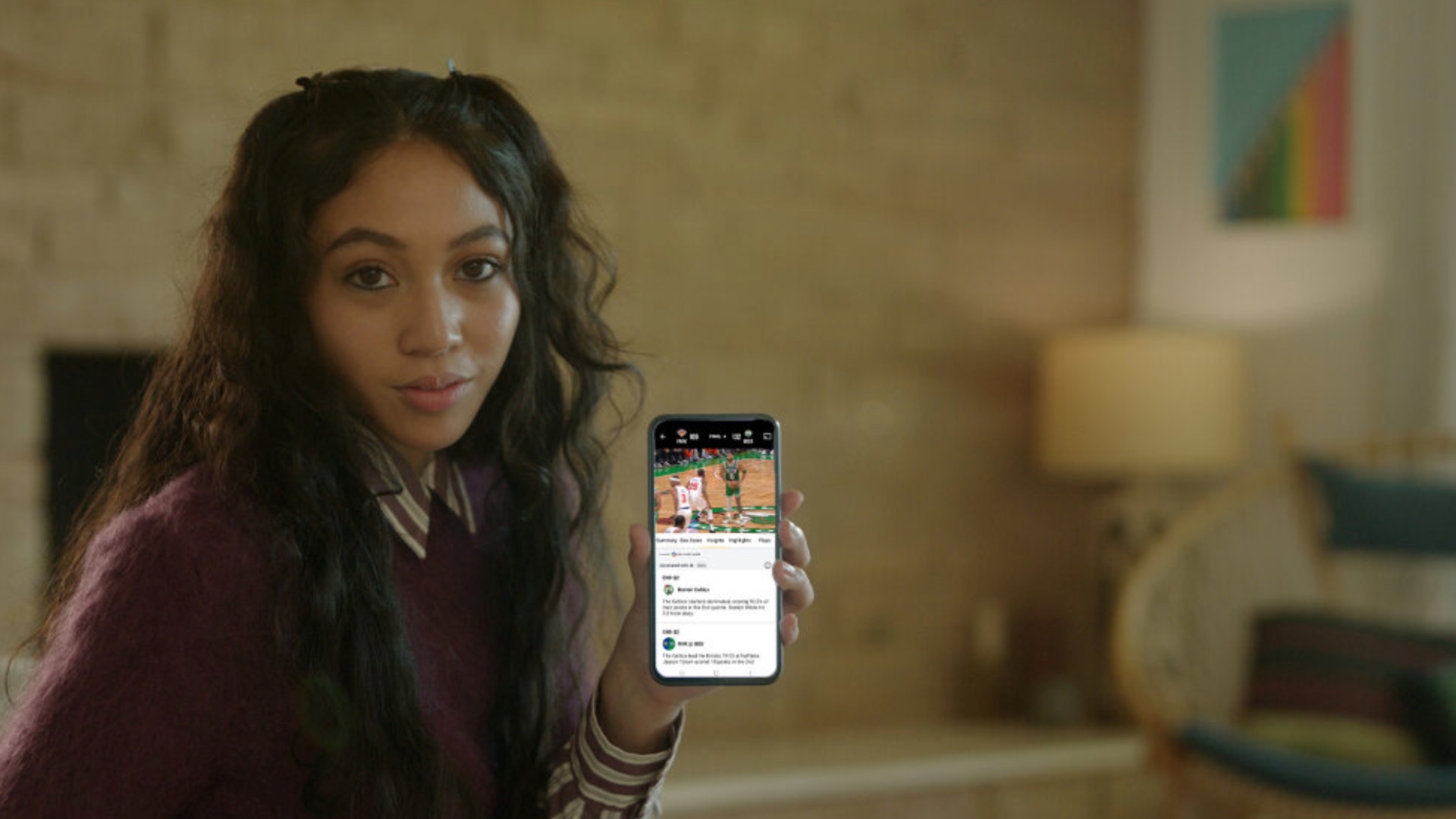 A person with long hair is indoors, holding up a smartphone displaying a social media page. The room has brick walls and a lamp in the background.