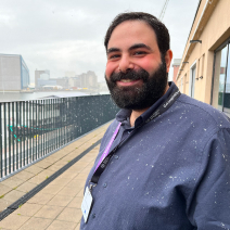 Man with a beard smiling outdoors on a balcony with light rain, wearing a blue shirt and a badge, city skyline in the background.