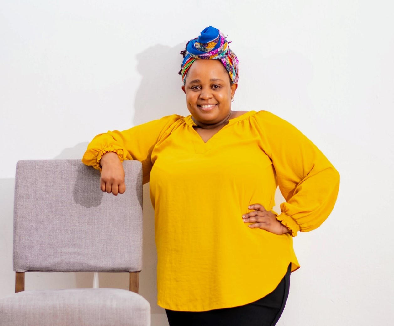 A confident woman in a yellow blouse and colorful headwrap stands with one hand resting on a gray chair against a white background.