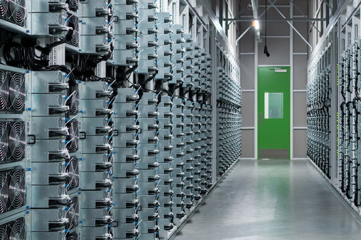 A modern data center with rows of server racks and cooling fans, illuminated by overhead lights, leading to a green exit door.