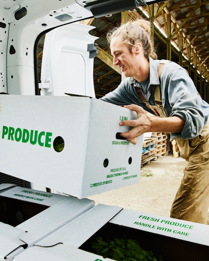 A man unloading a box labeled "produce" from a van inside a greenhouse.