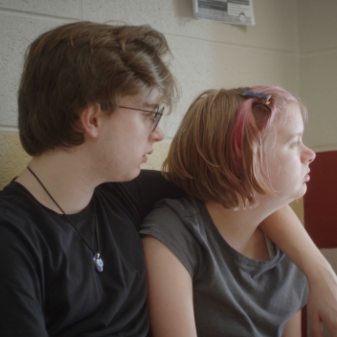 Two young adults sitting close together on a bench, looking thoughtful, one with pink hair and sunglasses on their head.