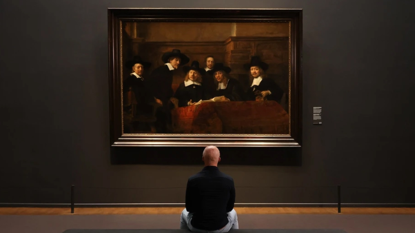 A man sitting on the floor, observing rembrandt's painting "the night watch" displayed on a museum wall.
