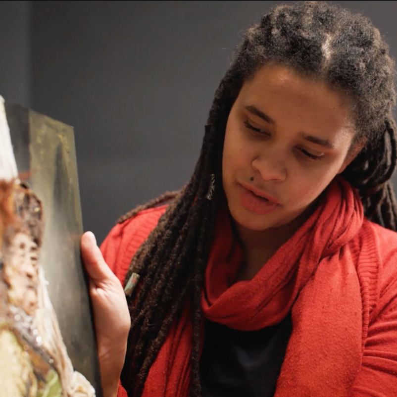 A woman with dreadlocks, wearing a red scarf, examines a painting closely in an indoor setting.