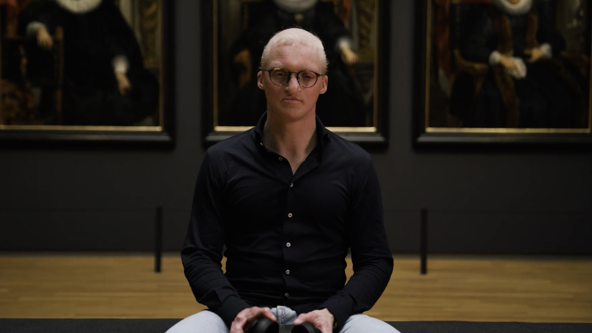 A man with pale skin and white hair meditating on the floor, sitting cross-legged in a gallery with classical paintings in the background.