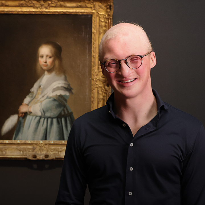 A smiling young man with glasses standing next to a framed painting of a young girl in an art gallery.
