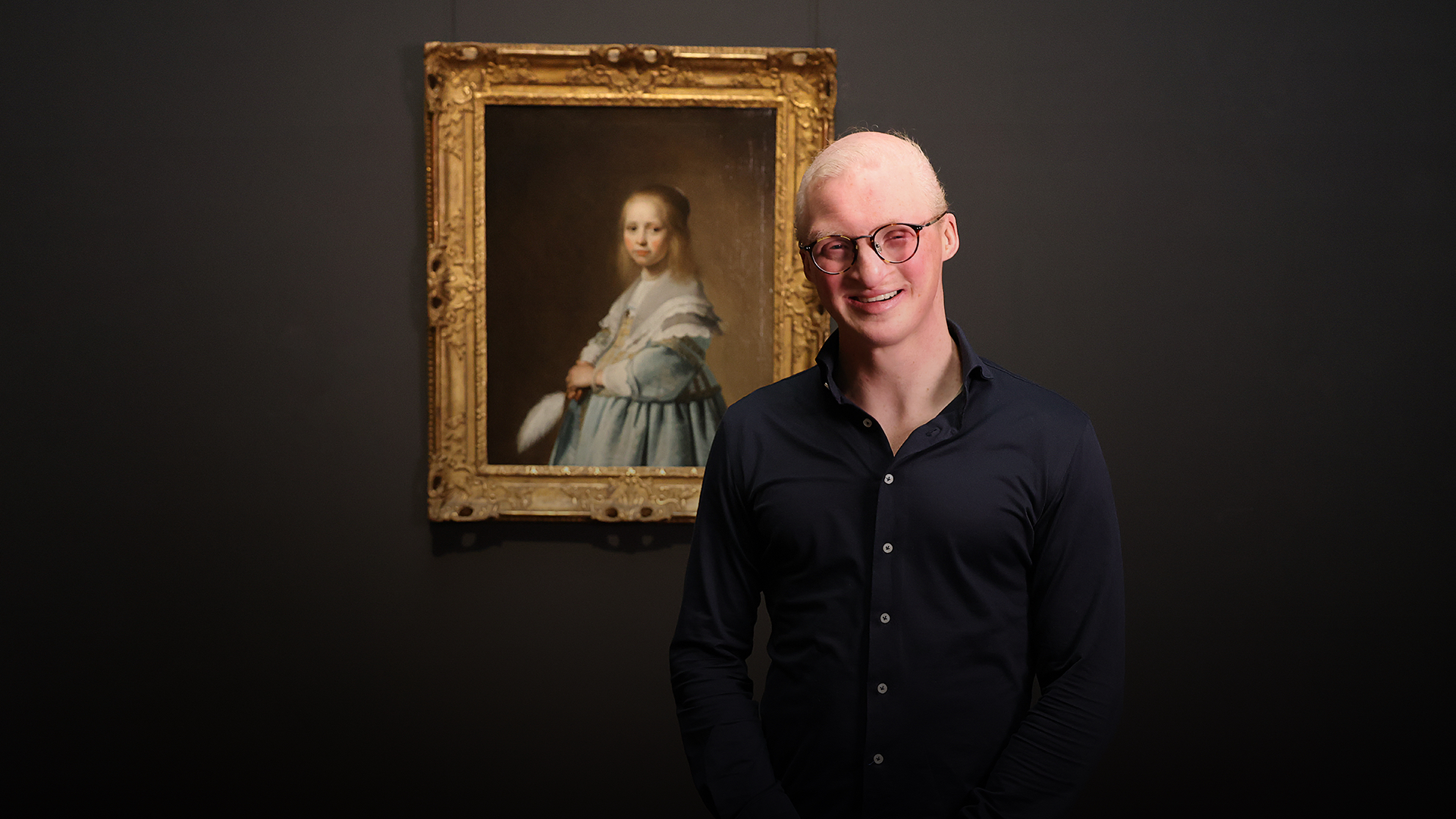 A man with glasses smiling while standing next to a painting of a girl in a blue dress, framed in gold, against a dark background.