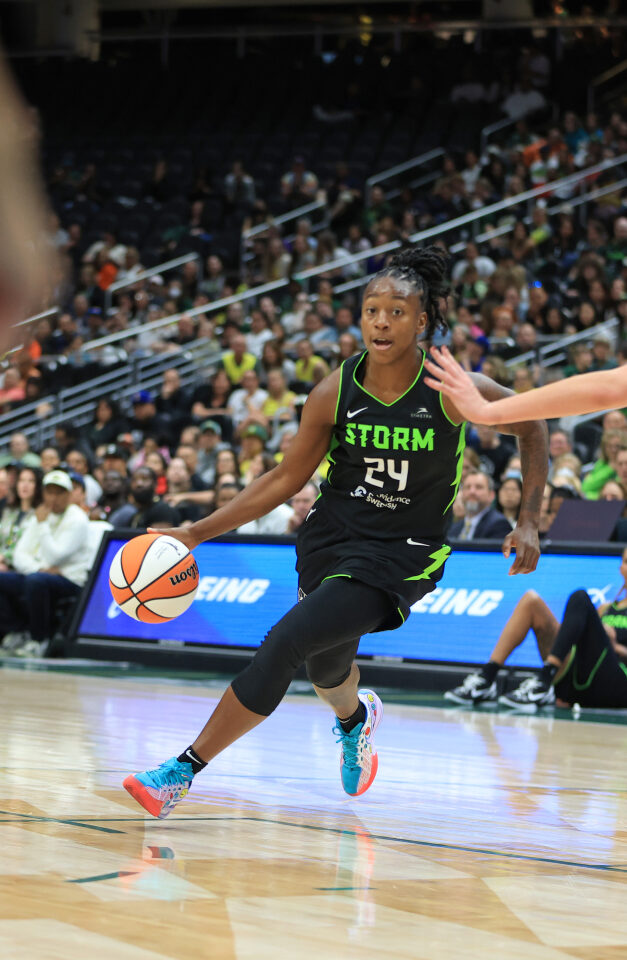 A basketball player wearing a black "Storm" jersey dribbles the ball on the court while a player defends her.