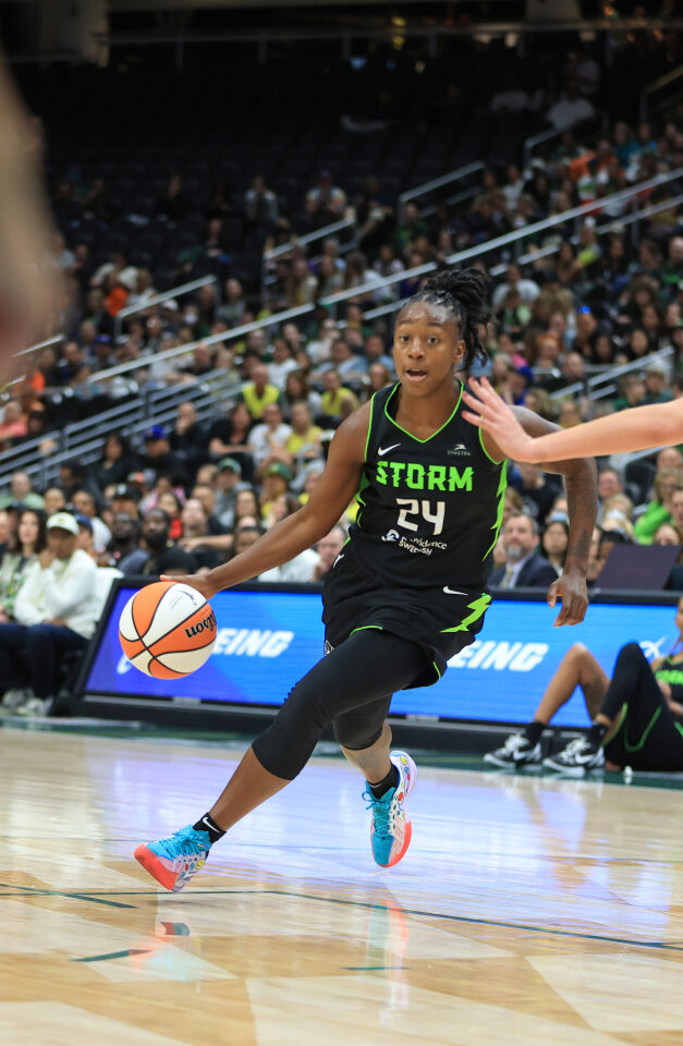 A basketball player wearing a black "Storm" jersey dribbles the ball on the court while a player in a white jersey defends her.