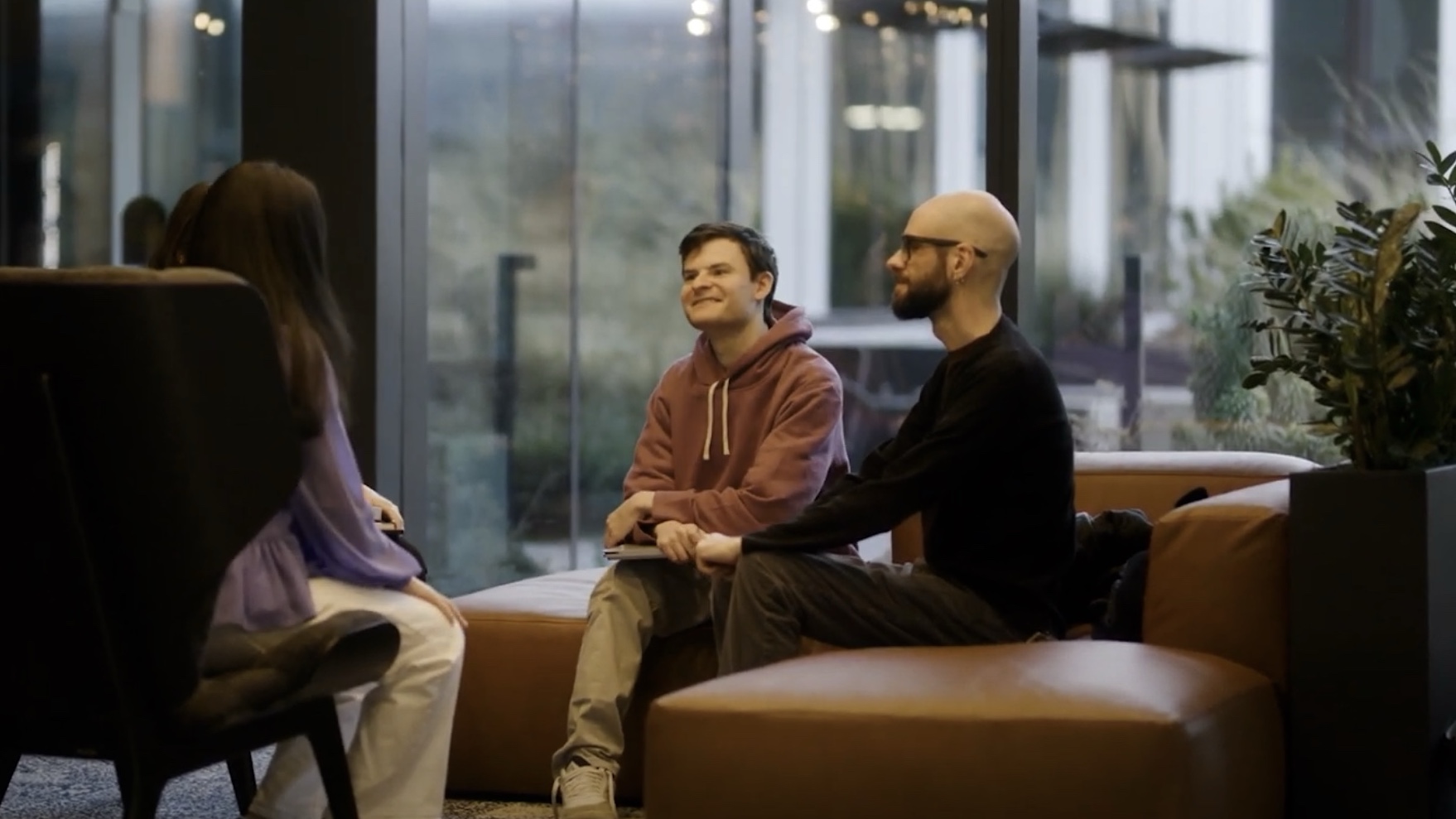 Three people sit on brown couches in a modern indoor space, engaged in conversation. Large windows reveal a blurred outdoor view in the background.