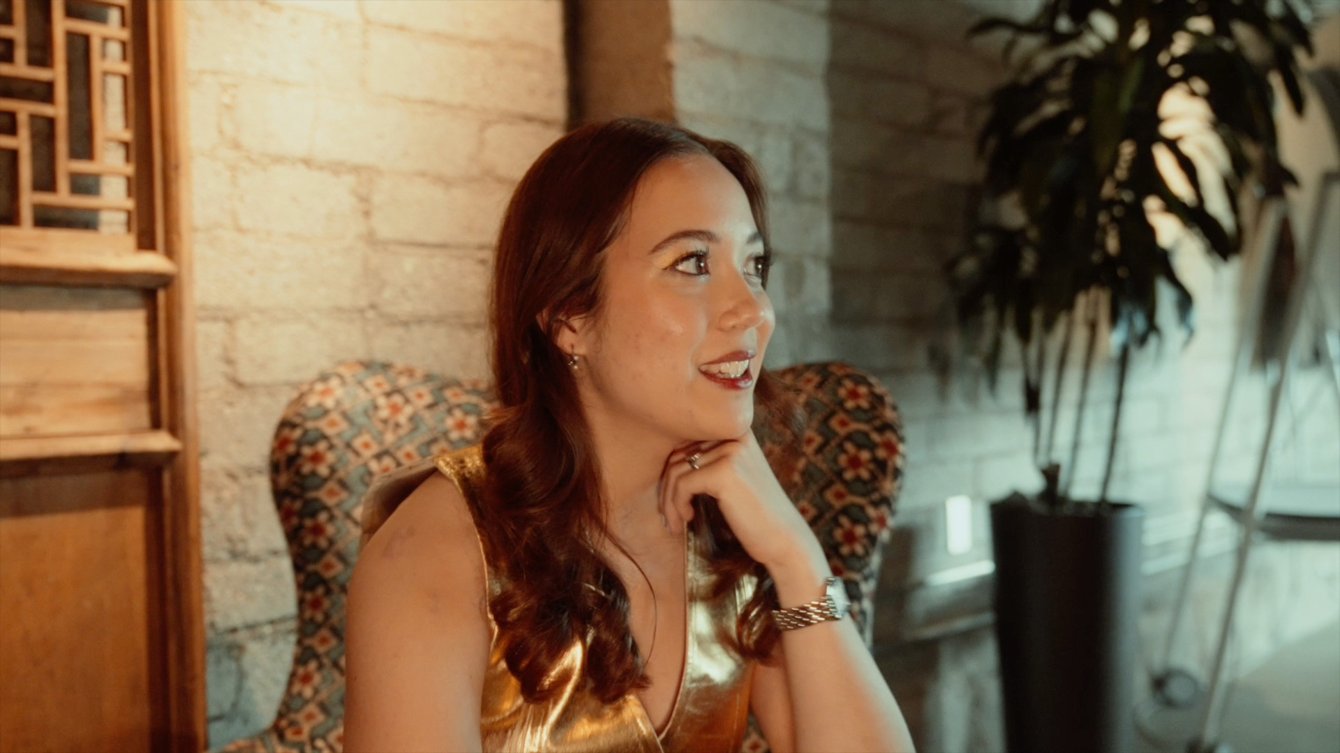 A woman with shoulder-length hair smiling, looking away from the camera, seated inside a warmly lit room with a brick wall and plants in the background.