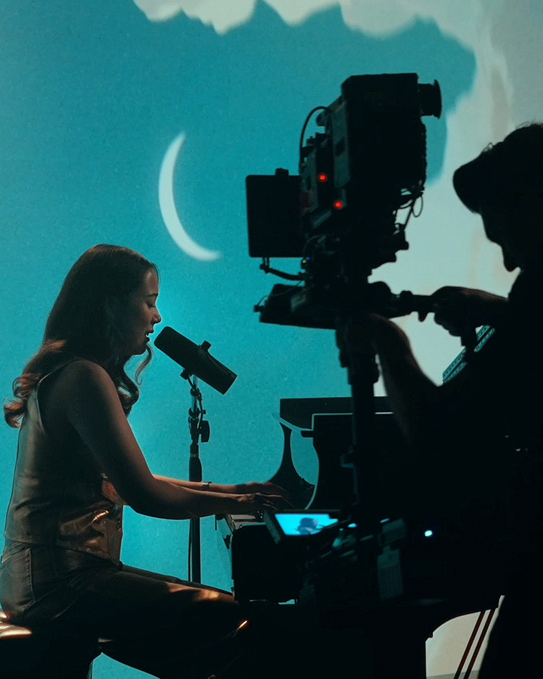 A woman sings into a microphone while playing piano on a set with a crescent moon backdrop, filmed by a nearby camera operator.