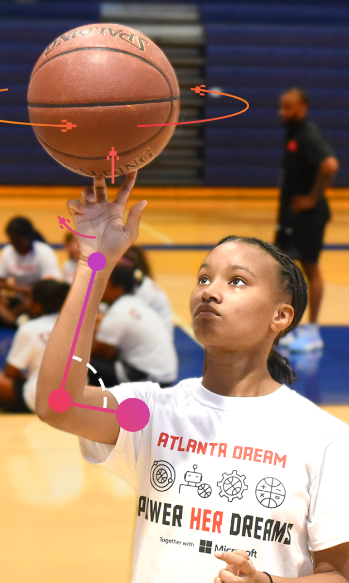 A young girl spins a basketball on her finger in a gym. She wears a white shirt with the text "Atlanta Dream Power Her Dreams." Other individuals and basketballs are in the background.