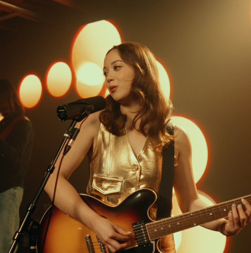 A female musician with a guitar singing into a microphone on a stage, illuminated by soft orange lights in the background.
