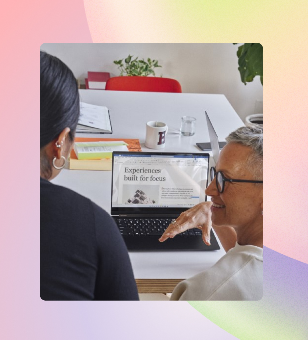 Two professionals, one with short gray hair and another with long black hair, are smiling and discussing a presentation on a laptop in a modern office setting.