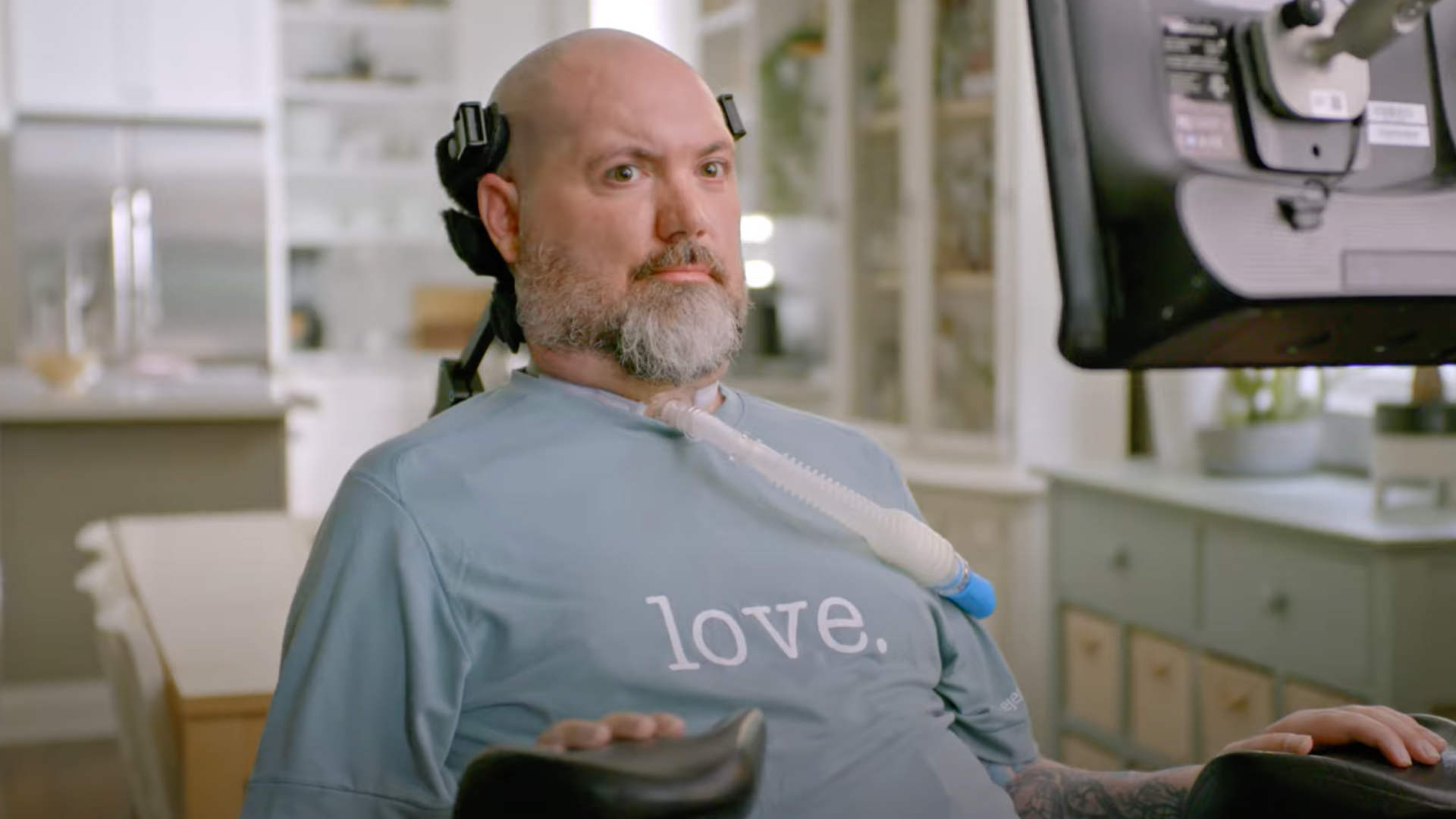 A bald man with a beard, using a ventilator and headgear, sits in a wheelchair with a computer screen in front of him.