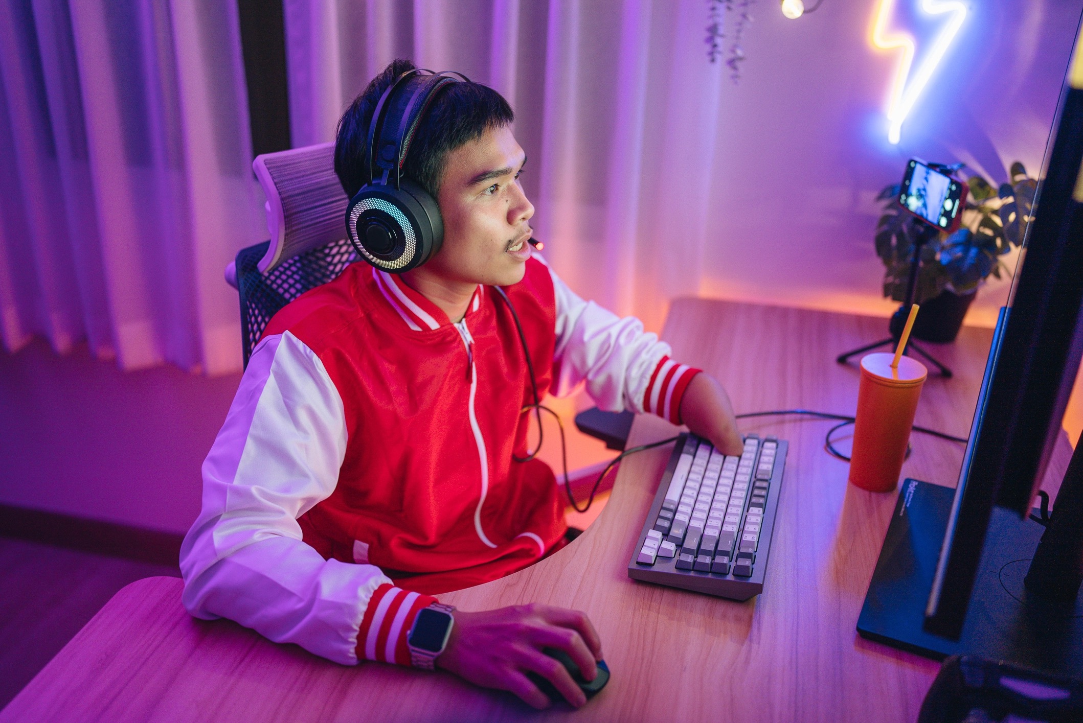 A young man wearing a red jacket and headset sits at a desk, using a computer with a keyboard and mouse, illuminated by colorful neon lights.