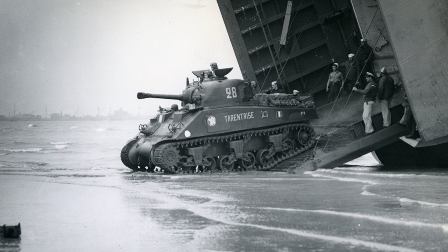 A military tank marked "28" exits a landing craft onto a beach, with several soldiers nearby.