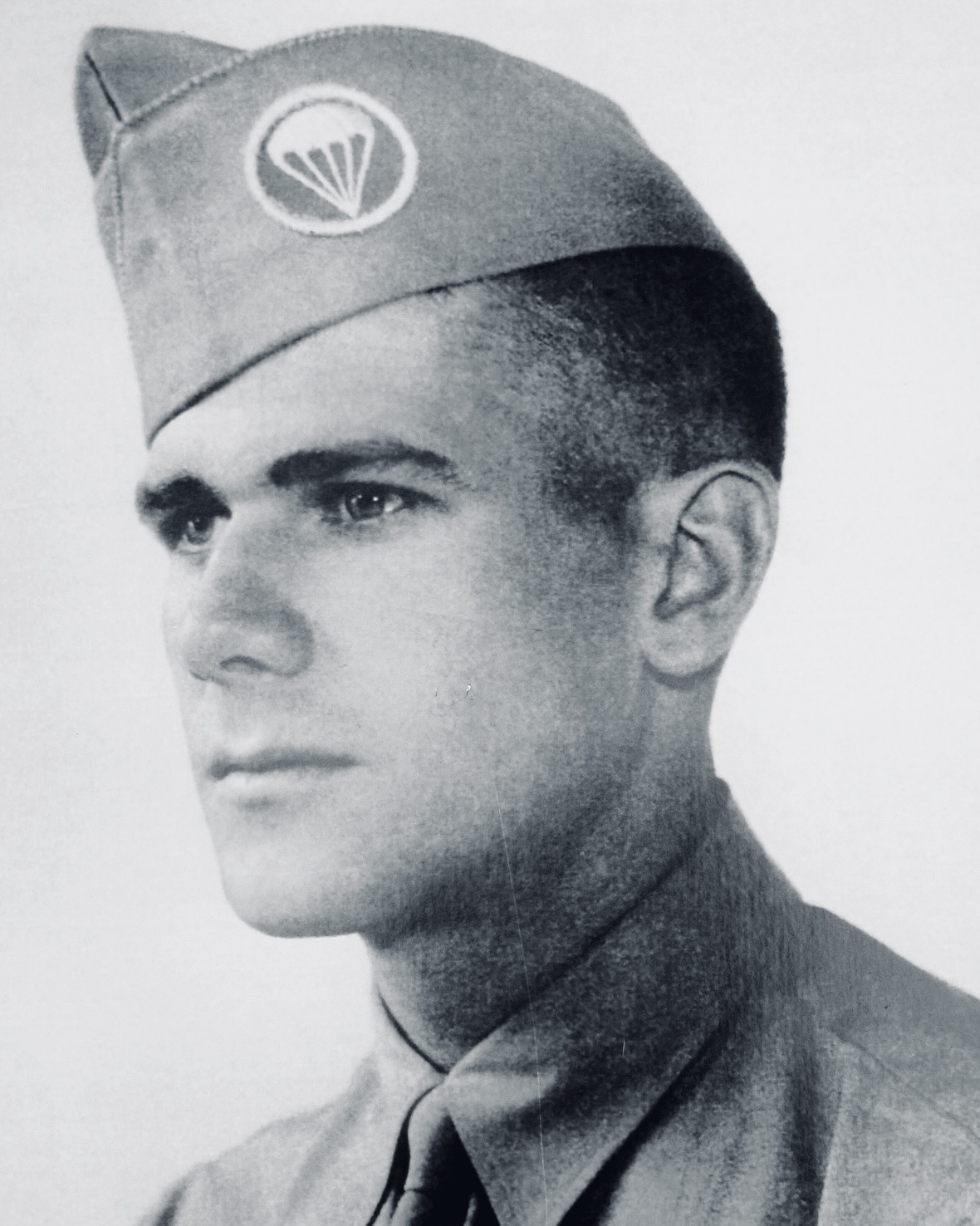Black-and-white portrait of a man in a military uniform wearing a cap with a parachute insignia, looking to the side.