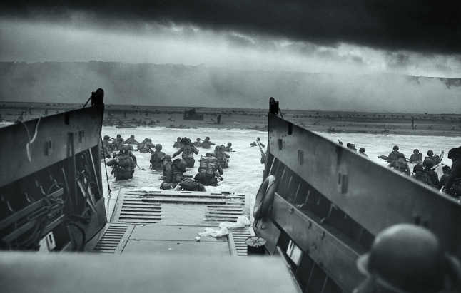 Black and white photo of soldiers wading through water after disembarking from a landing craft onto a beach under dark, cloudy skies.