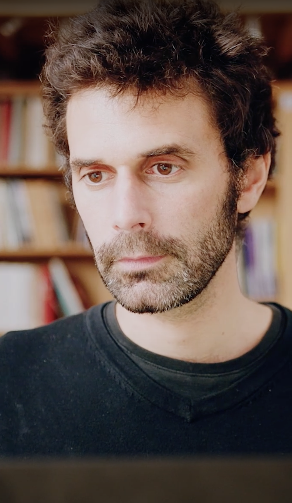 A man with a beard and wavy hair wearing a black sweater is looking ahead with a serious expression. Bookshelves with books are blurred in the background.