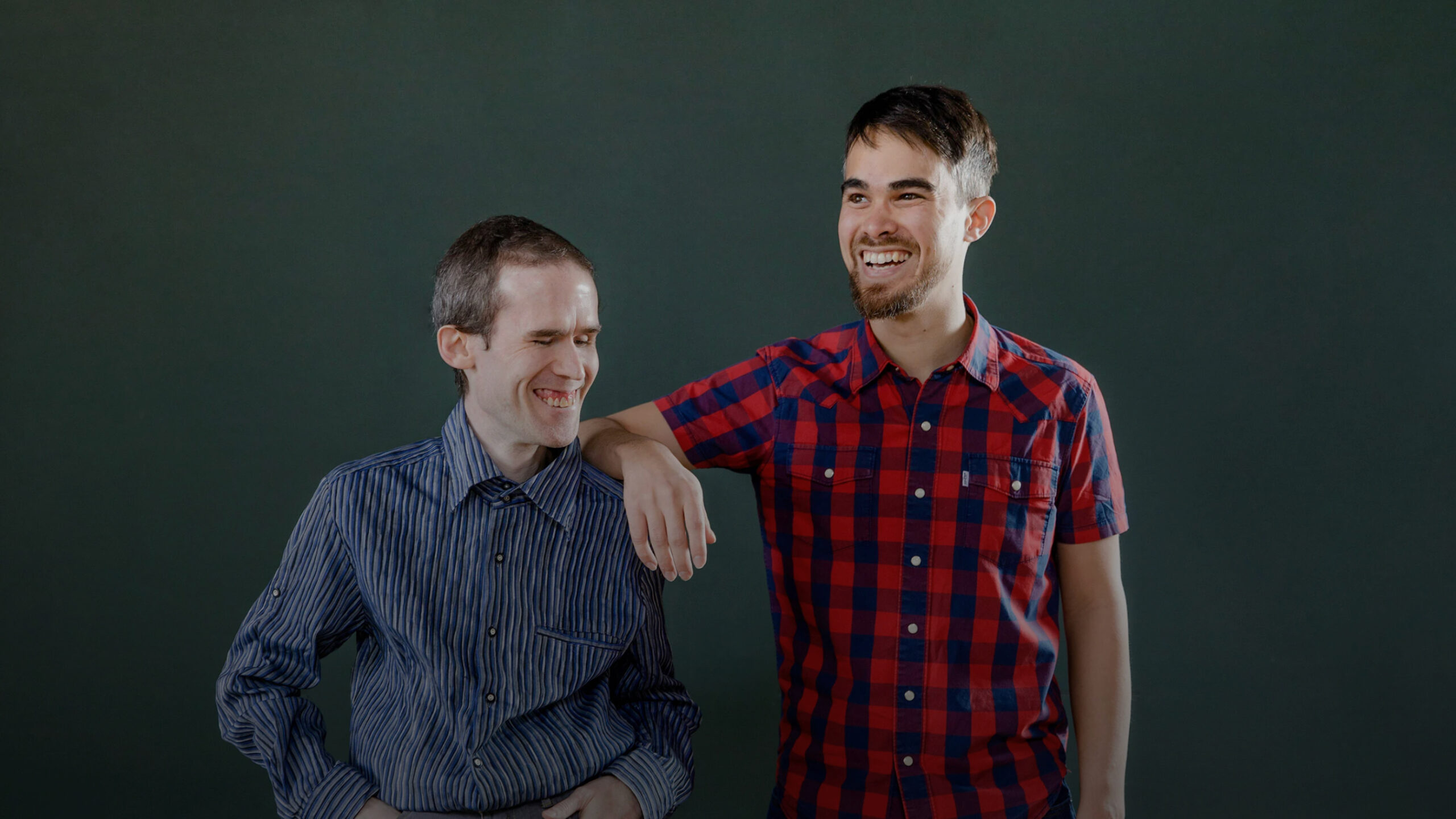 Two men smiling, one with his arm around the other, standing against a dark green background. they are wearing casual shirts.