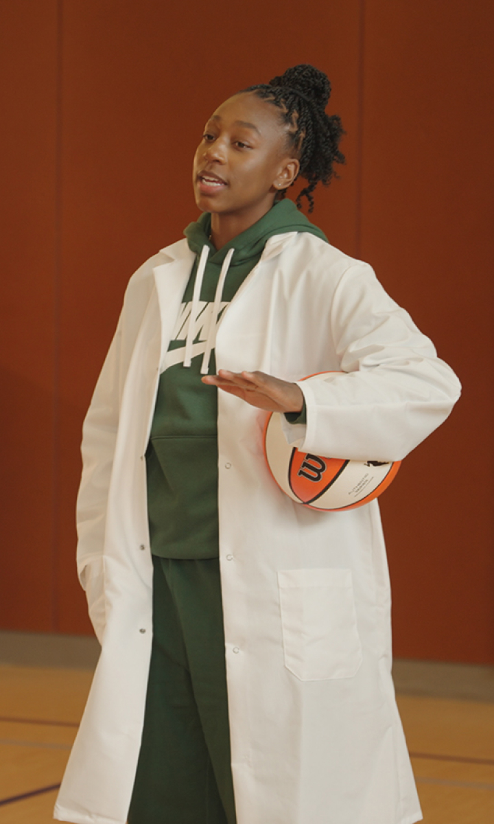 A young athlete in a white lab coat and green sports uniform holds a basketball under one arm in a gymnasium.