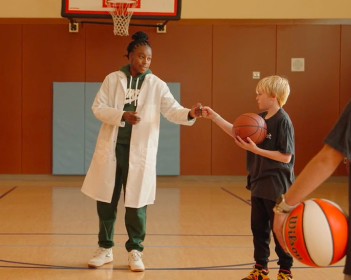A woman in a lab coat hands a basketball to a young boy in a gymnasium, with another basketball nearby.