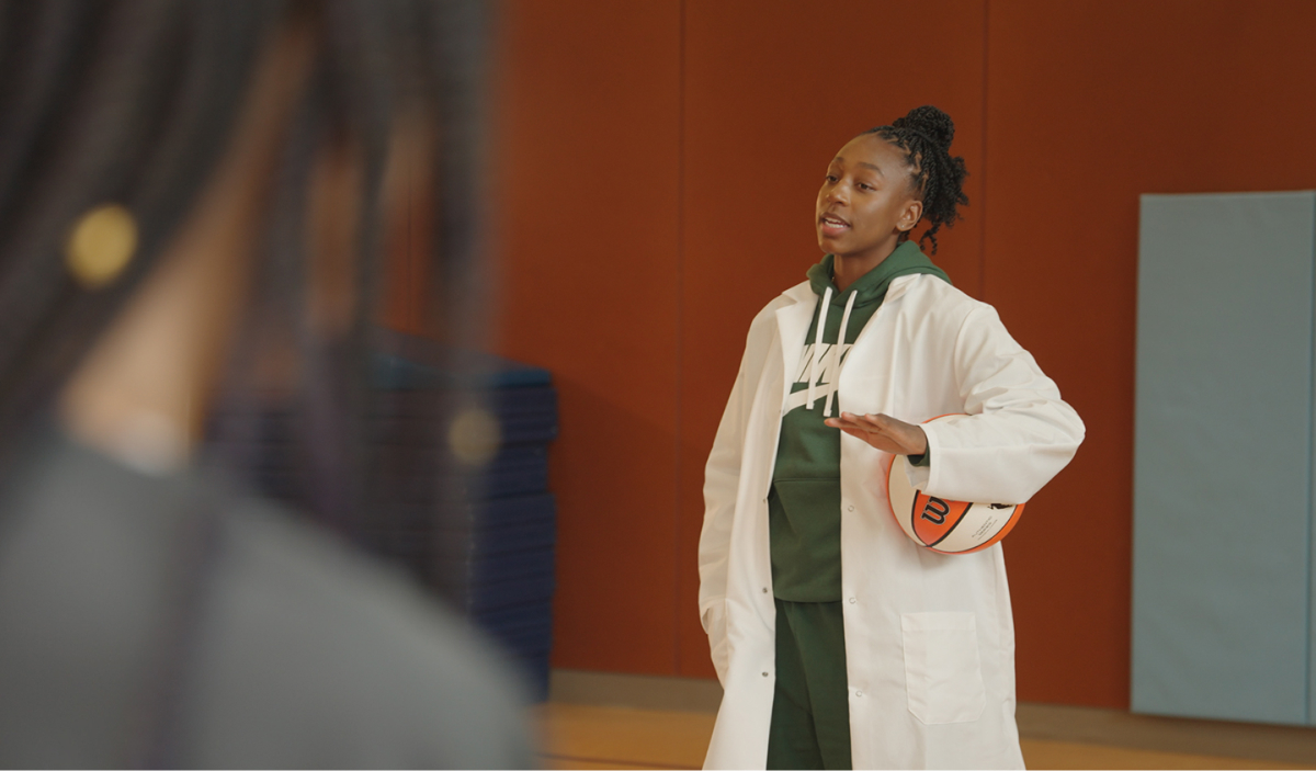 A young black woman in a lab coat speaking to a colleague in a classroom-like setting with a cheerful expression.