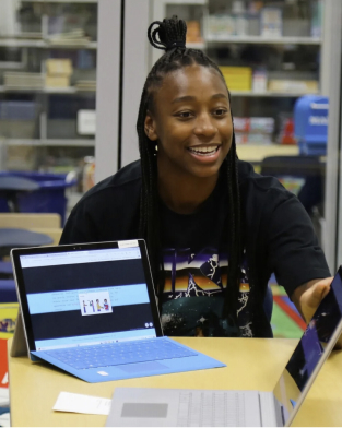 Young woman with a laptop in a library, smiling, wearing a black t-shirt, with her hair styled in a top bun.