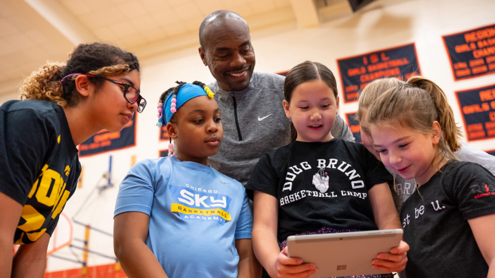 A group of four young girls and an adult male smiling as they look at a tablet together in a gymnasium.