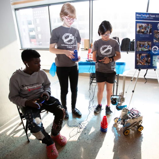 Three teenagers in matching t-shirts operate robotic devices in a classroom setting, with a poster about the blue twilight team 22 in the background.