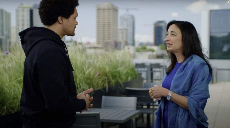 Two people having a conversation on an outdoor terrace with city buildings in the background. The man is wearing a black hoodie and the woman is in a blue shawl.