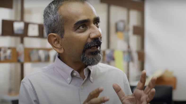 A man with short gray hair and a beard, wearing a light-colored shirt, gestures with his hands while speaking indoors. The background includes blurred wall decorations and light coming from the side.