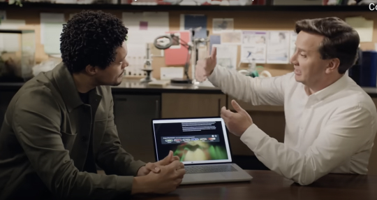 Two men sitting at a table in an office setting, engaged in discussion. One man gestures with his hand while the other listens attentively. A laptop displaying a chart is open on the table.