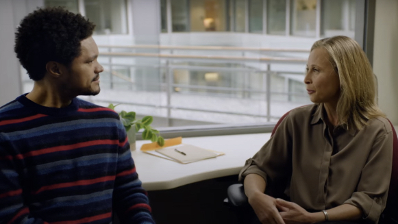 A man in a striped sweater and a woman in a brown blouse sit in an office, facing each other and having a conversation. A window and a potted plant are visible in the background.