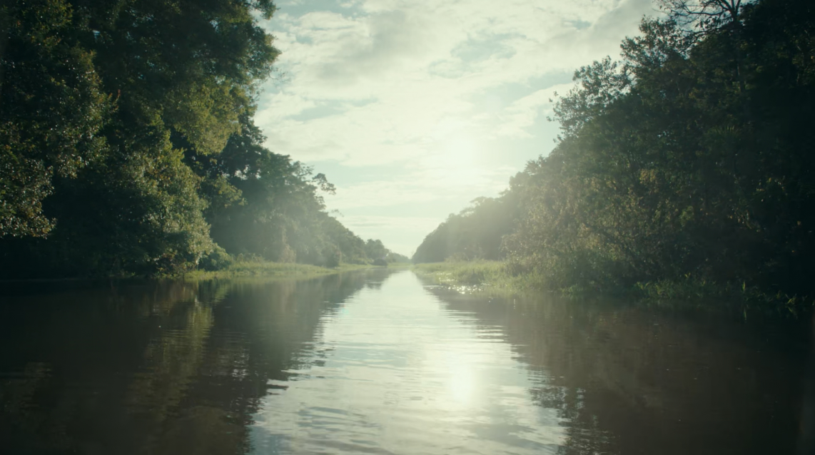 A calm river flows between lush, green trees under a partly cloudy sky with the sun shining in the background.