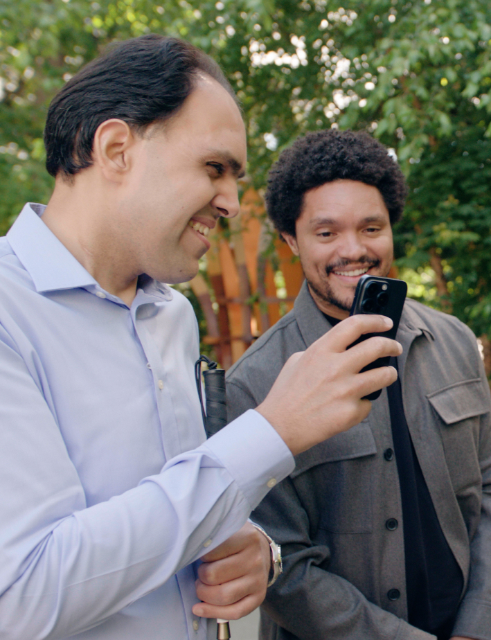 Two men smiling and looking at a smartphone outside, with greenery in the background.