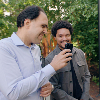 Two men smiling while looking at a mobile phone outdoors. One man is wearing a light blue shirt and holding a cane, and the other is in a grey jacket standing beside him. Trees are visible in the background.