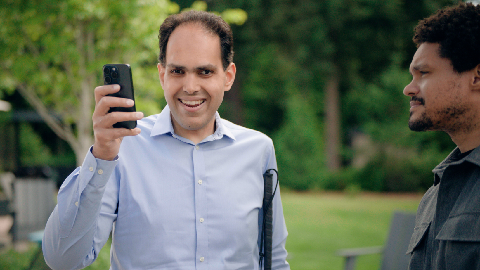 A person holding a smartphone smiles at the camera while another individual stands nearby outdoors. They are surrounded by greenery.