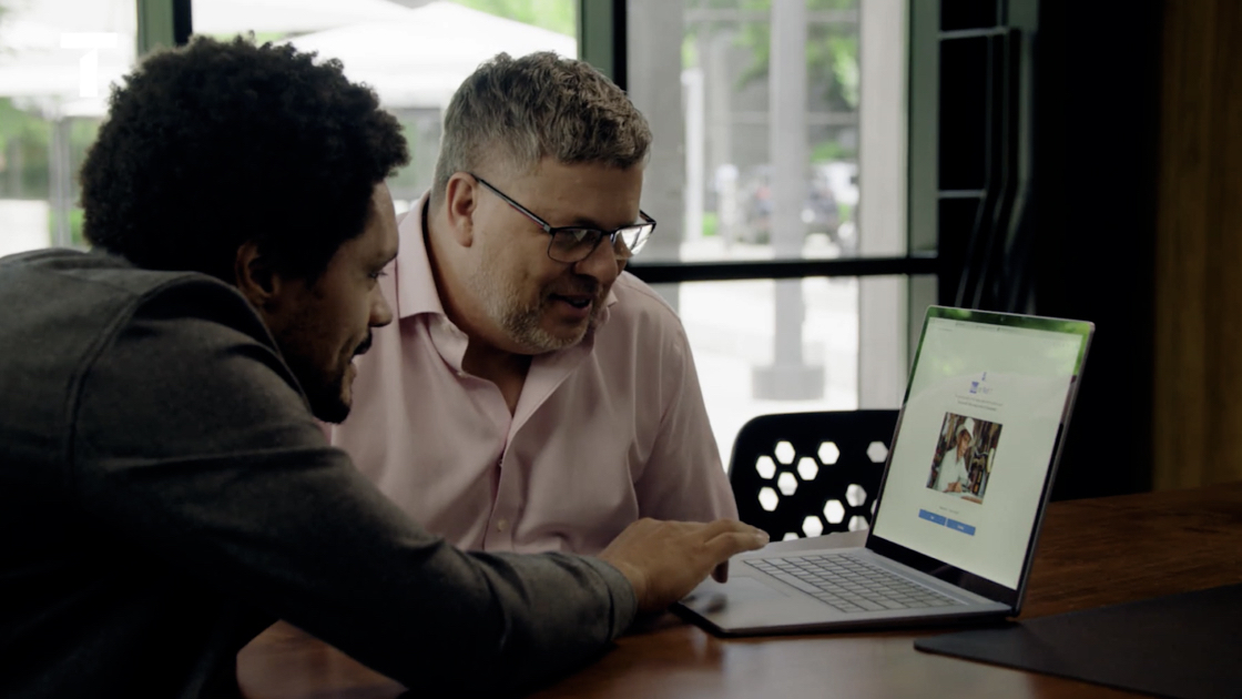 Two men sitting at a table look at a laptop screen together. One points at the screen while the other attentively watches. An outdoor scene is visible through the window in the background.