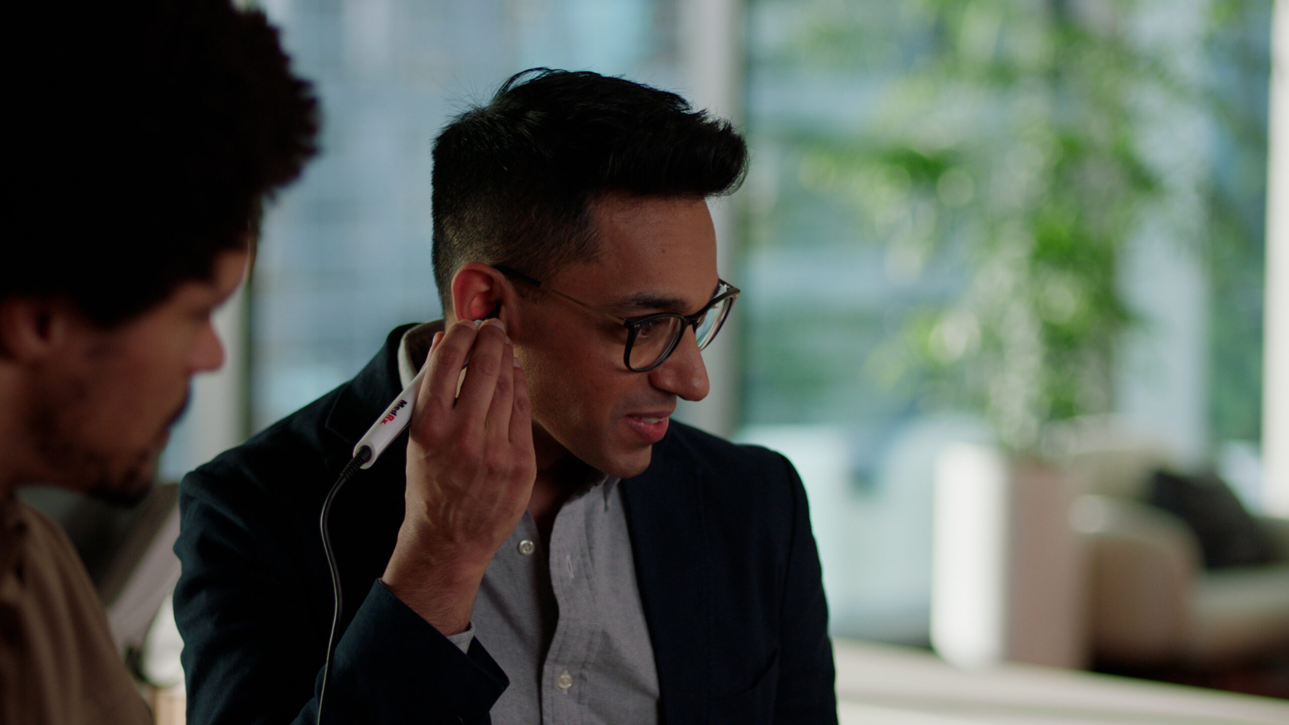 A man in glasses and a blazer holds a white hearing aid device to his ear, while another person stands nearby in an indoor setting.