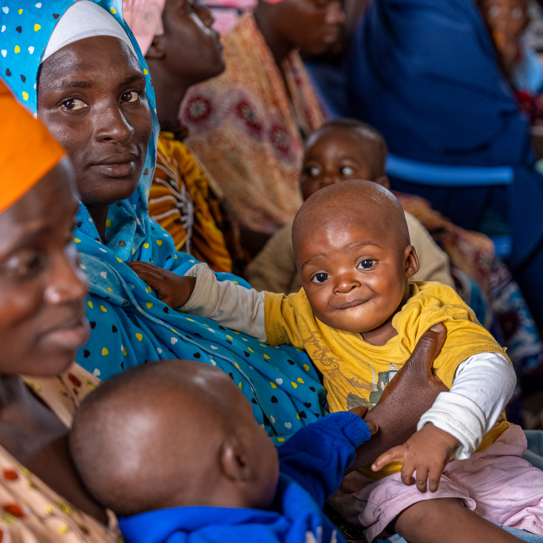 Adults and children seated closely in a crowded indoor setting. An adult in blue patterned attire holds an infant wearing a yellow shirt, while another child in blue is partially visible in the foreground.