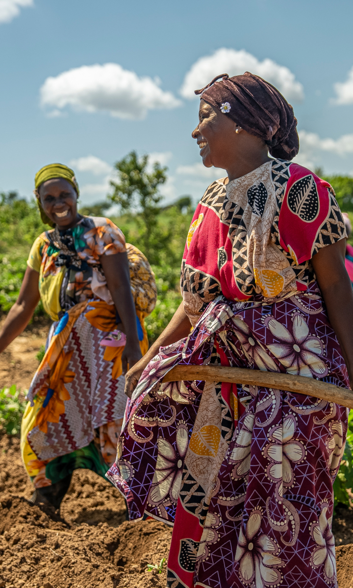 Two women in brightly colored attire work together in a sunny field, one holding a hoe, both smiling and appearing to enjoy their tasks.